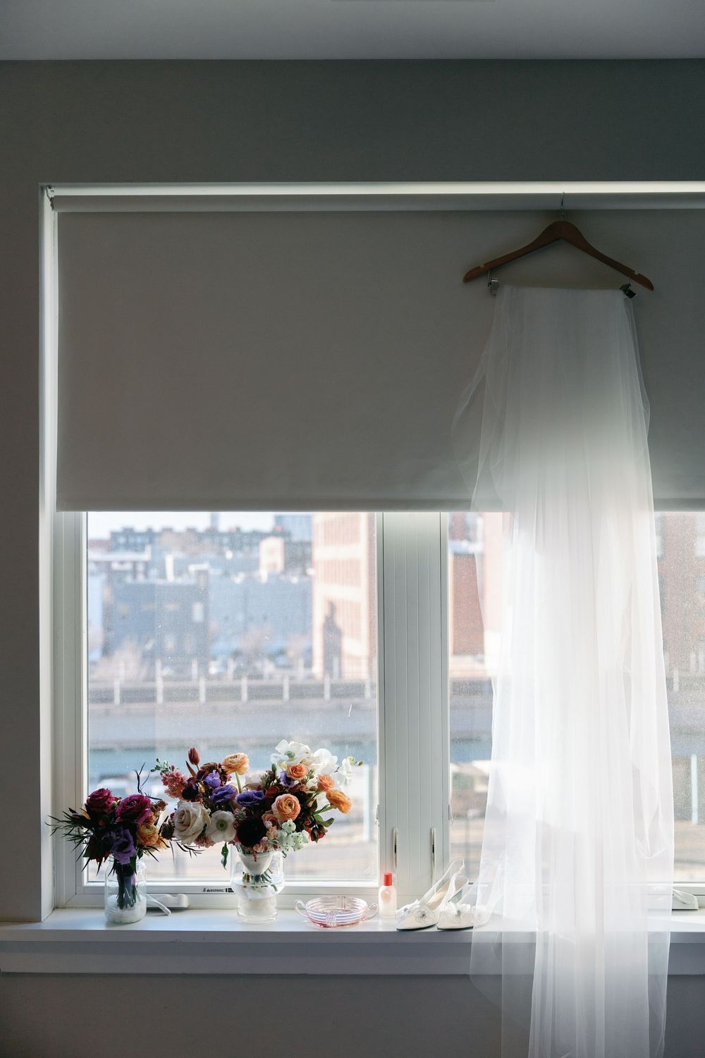 Wedding veil hanging by window with floral arrangements and city view.