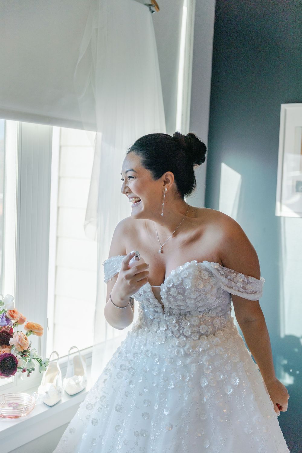 A joyful bride wearing an off-shoulder white wedding dress with floral appliques laughs near a window.