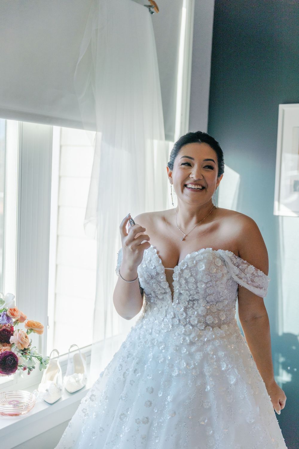 Bride in lace off-shoulder wedding gown by window with floral decorations.