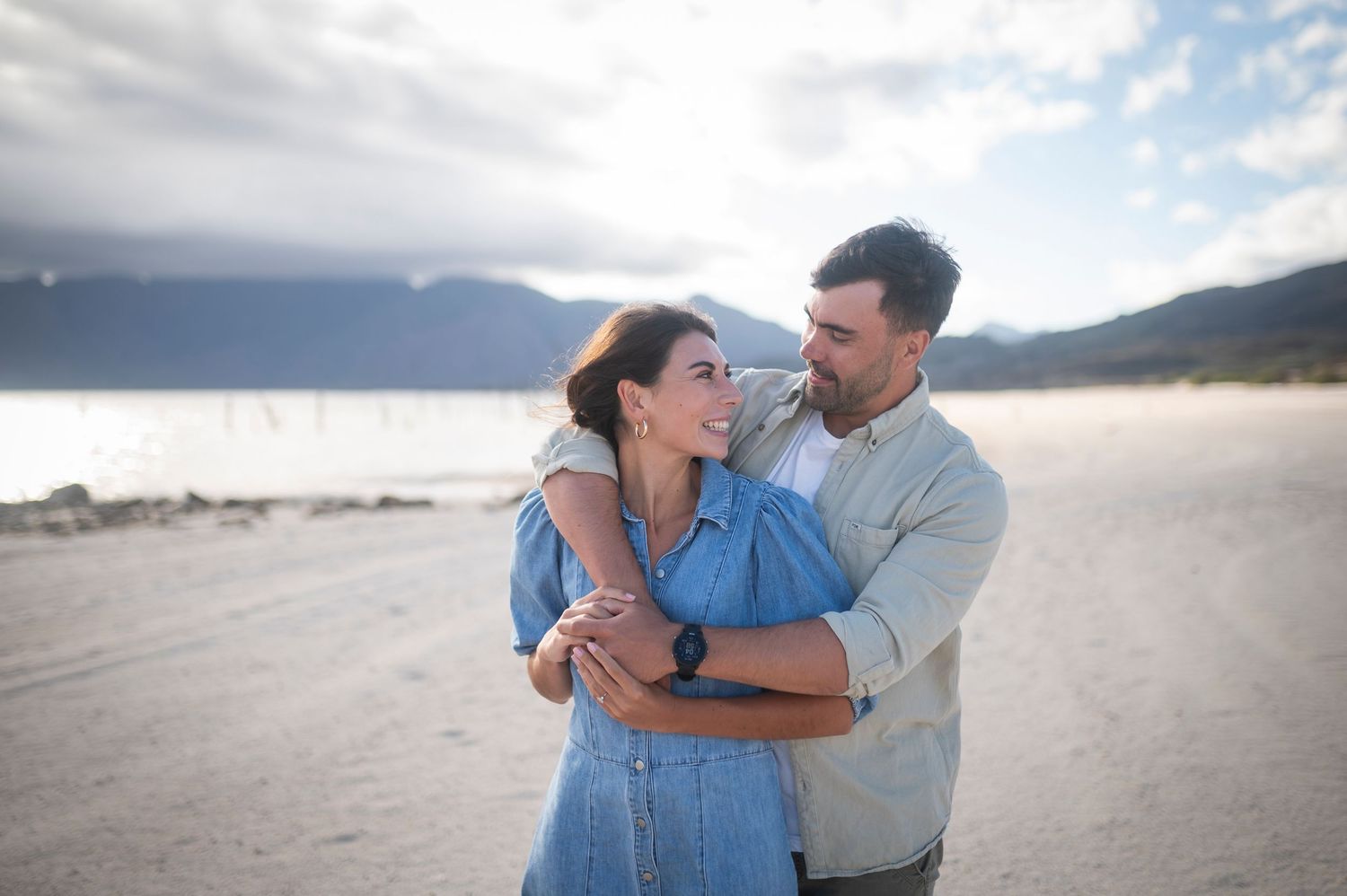 Romantic couple embraces and laughs together on a scenic beach with mountains and cloudy skies in the background.