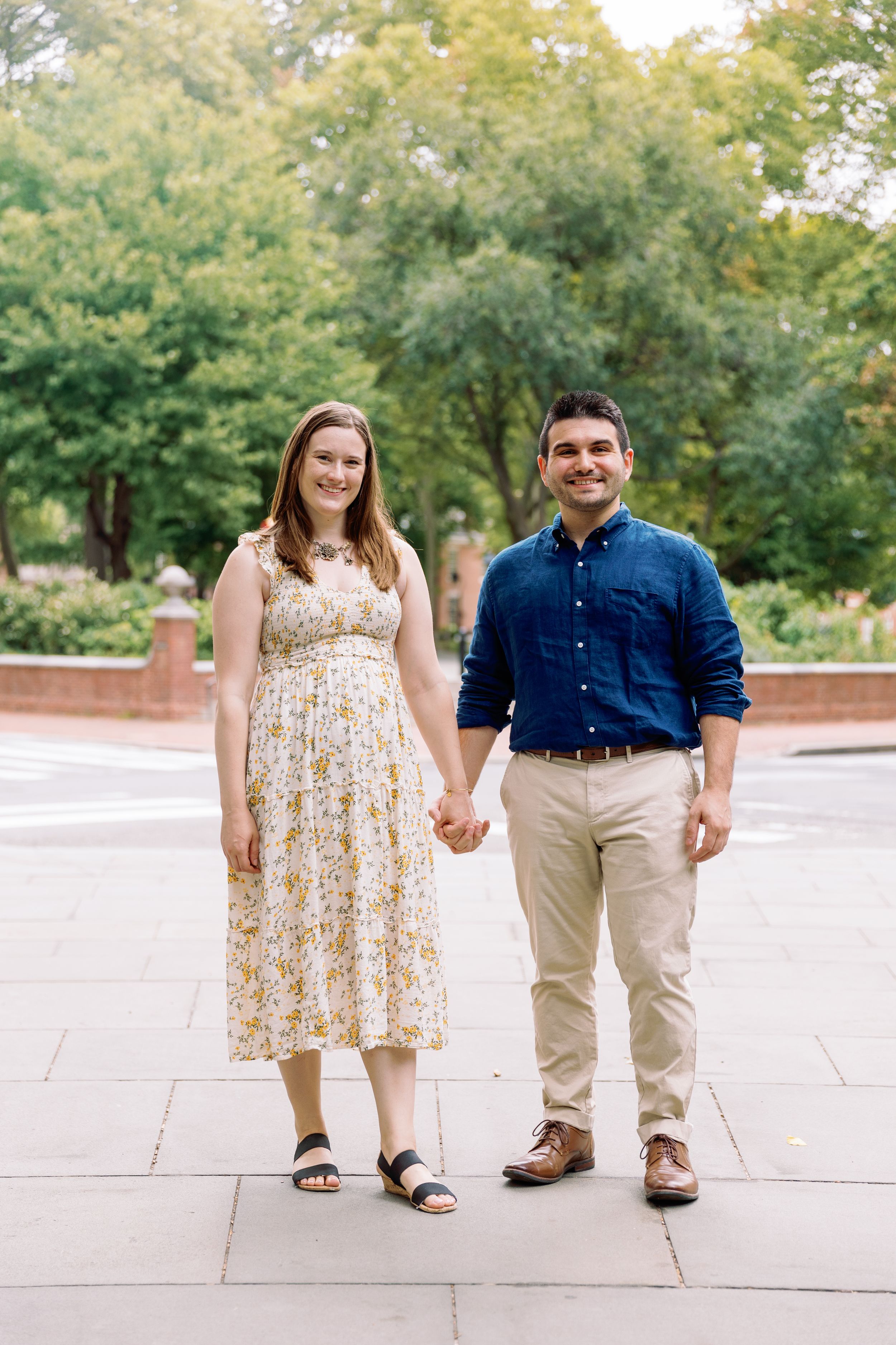 A couple holding hands walks together on a brick pathway surrounded by green trees.