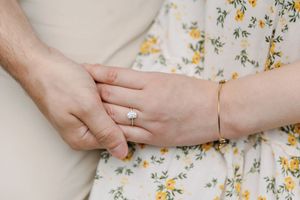 Two hands gently touch over a dress with yellow floral print, showcasing an engagement ring and delicate gold bracelet.