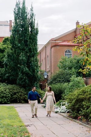 A romantic couple strolls along a stone pathway surrounded by lush greenery outside a brick building on a summer day.