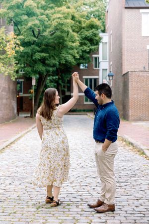A couple dancing together on a cobblestone street with brick buildings and green trees in the background.