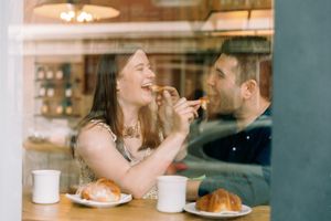 Couple shares a joyful moment while eating croissants and drinking coffee at a cozy cafe.