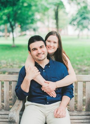 A couple embraces while sitting together on a wooden park bench surrounded by green trees and grass.