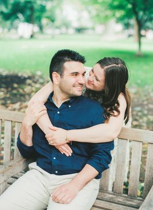 A couple shares an intimate moment while sitting together on a wooden park bench surrounded by green trees and grass.