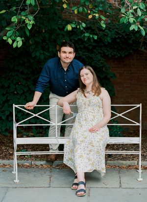 A couple poses together on a white metal garden bench against a brick wall with green foliage.