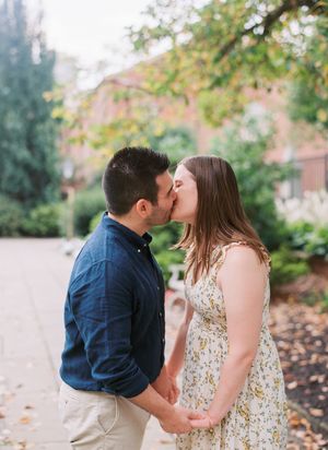 Two figures stand close together dancing outdoors in a landscaped garden area with trees in the background.