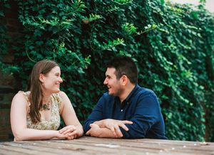 A couple sits at a wooden table outdoors enjoying conversation with lush green foliage in the background.