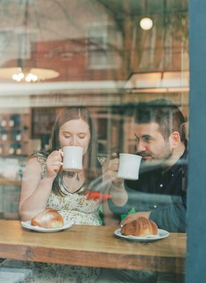 Through a glass window, two people share coffee and croissants at a cafe table in warm lighting.