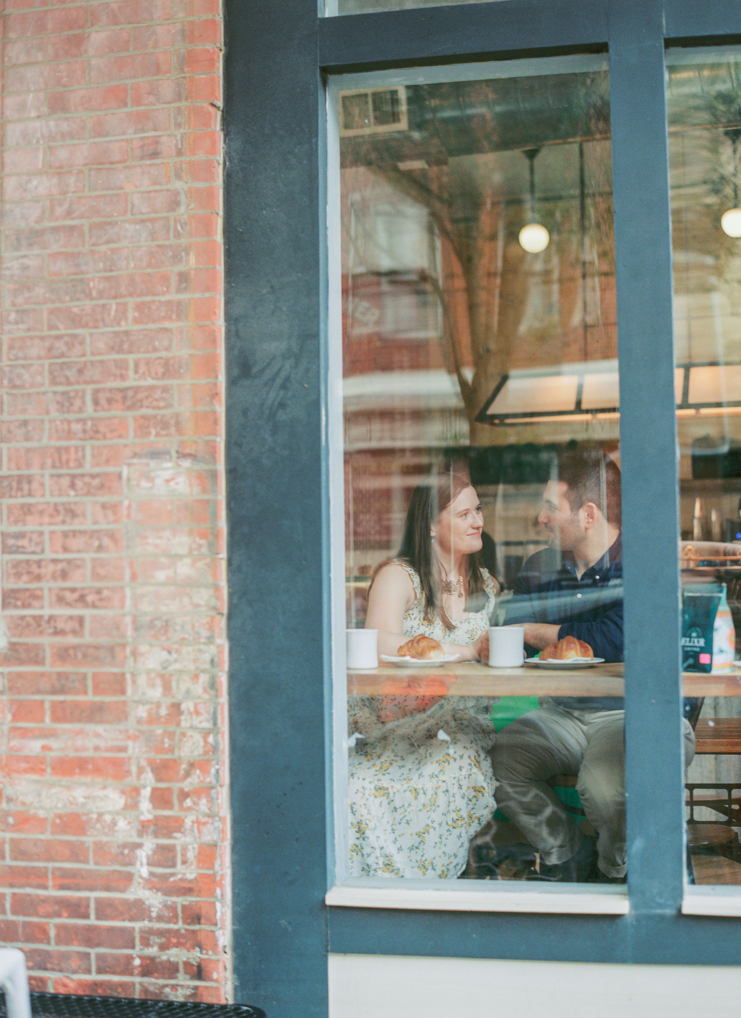 Two people enjoying coffee and conversation by a cafe window with exposed brick walls and natural lighting.