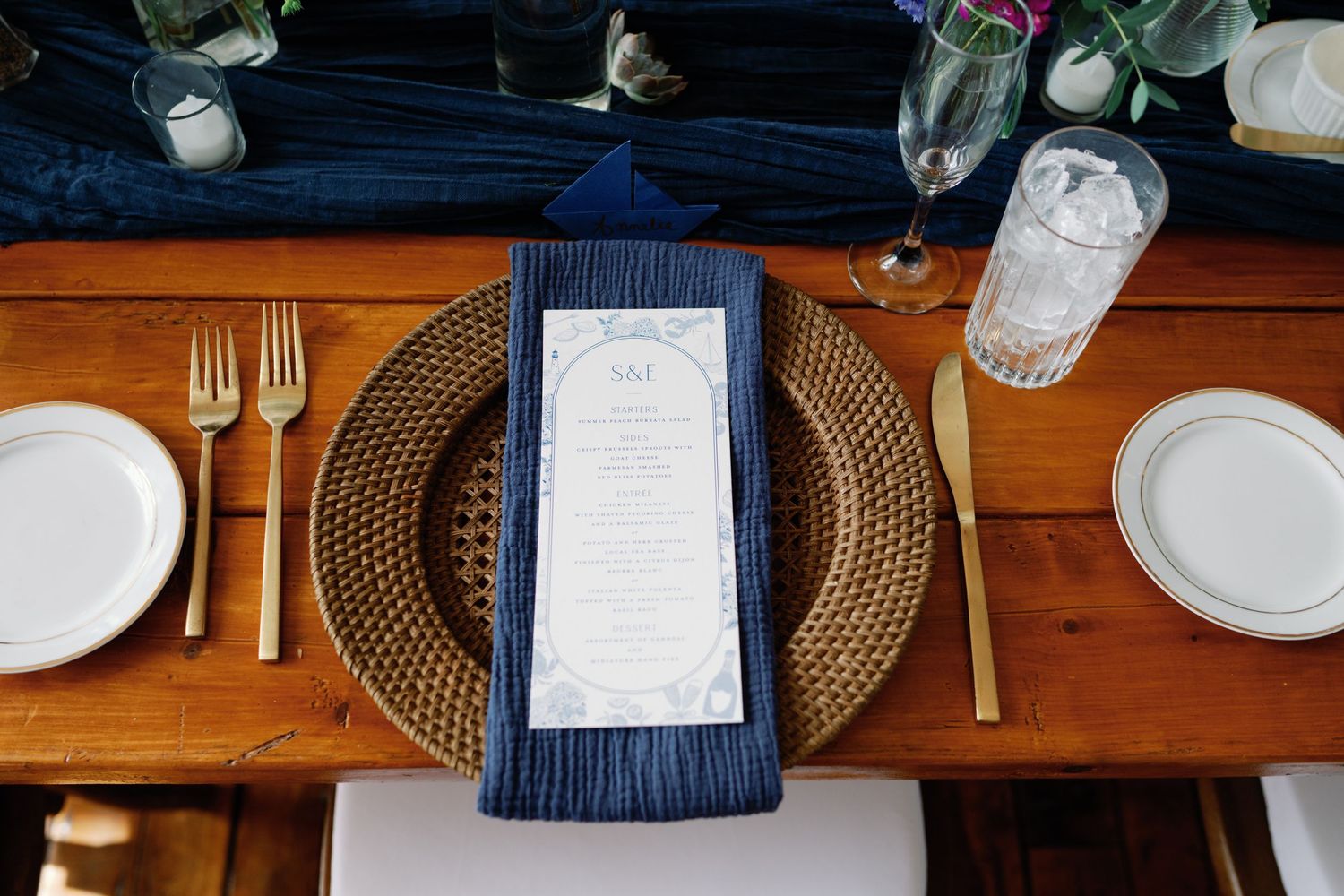 Elegant table setting with gold charger plate, navy blue napkin, and white menu card on wooden dining table.