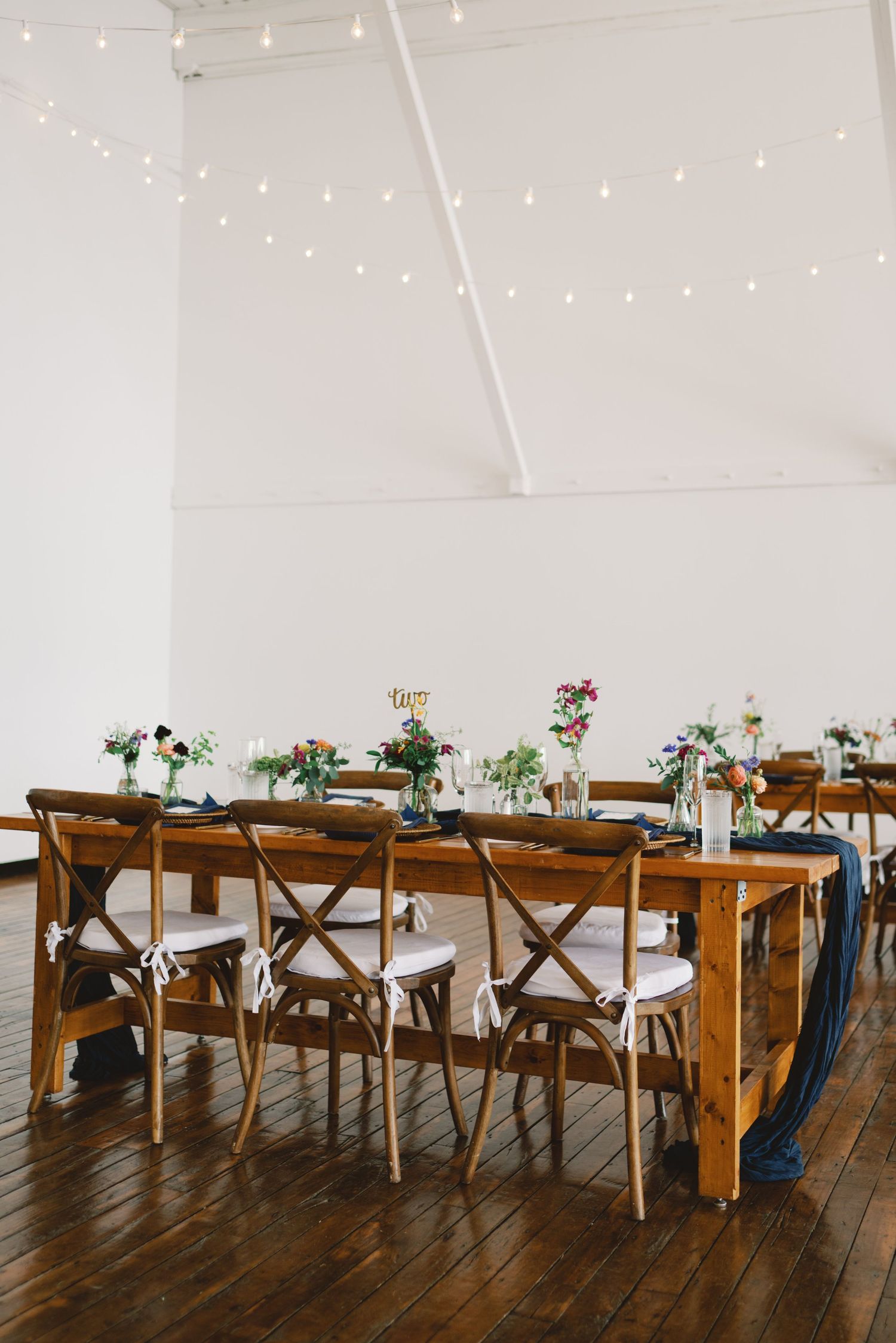 Long wooden farmhouse tables with cross-back chairs set up for wedding reception under twinkle lights on rustic plank floors.