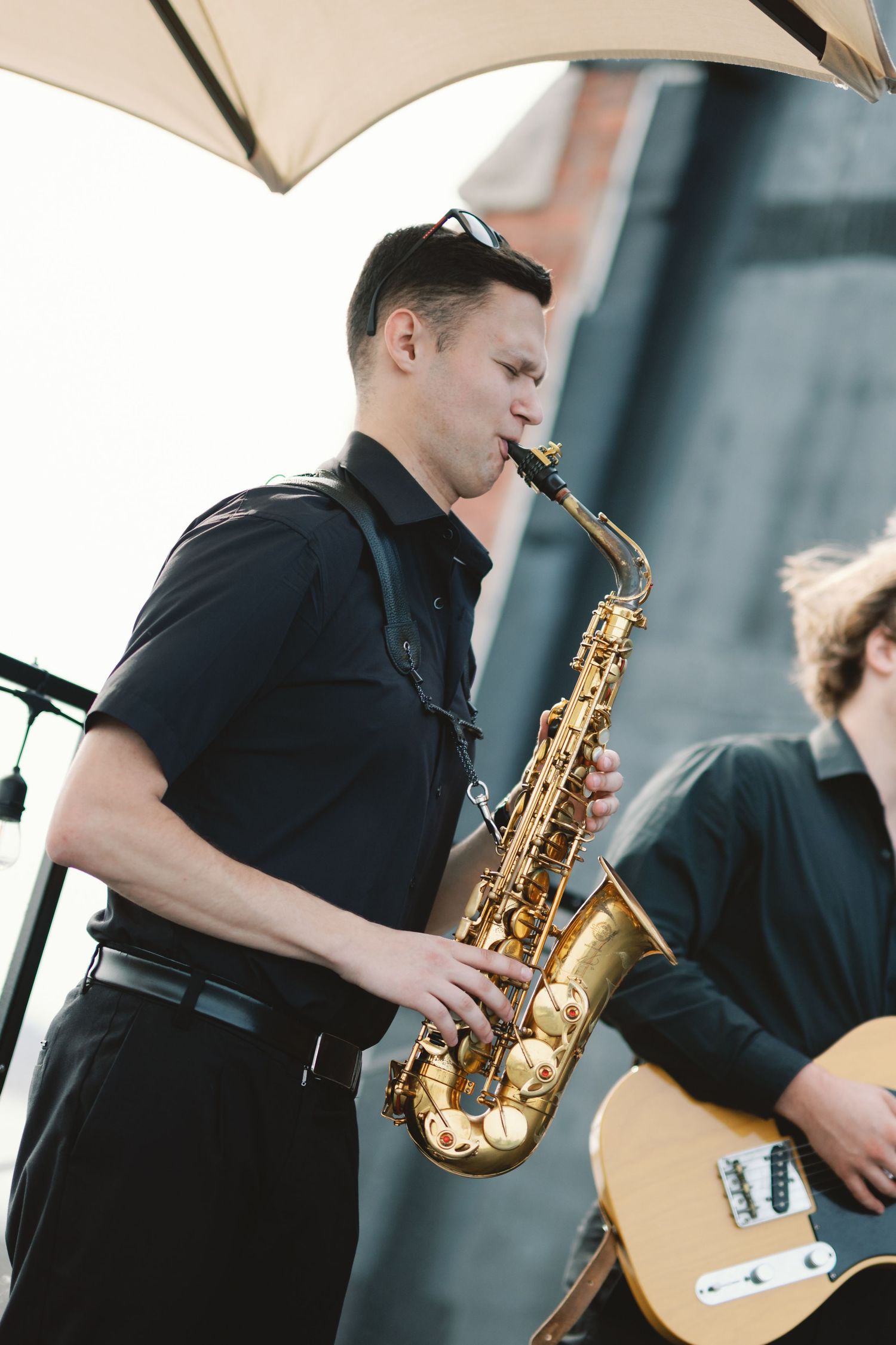 Musician plays golden saxophone under a beige umbrella while performing outdoors with guitarist in background.