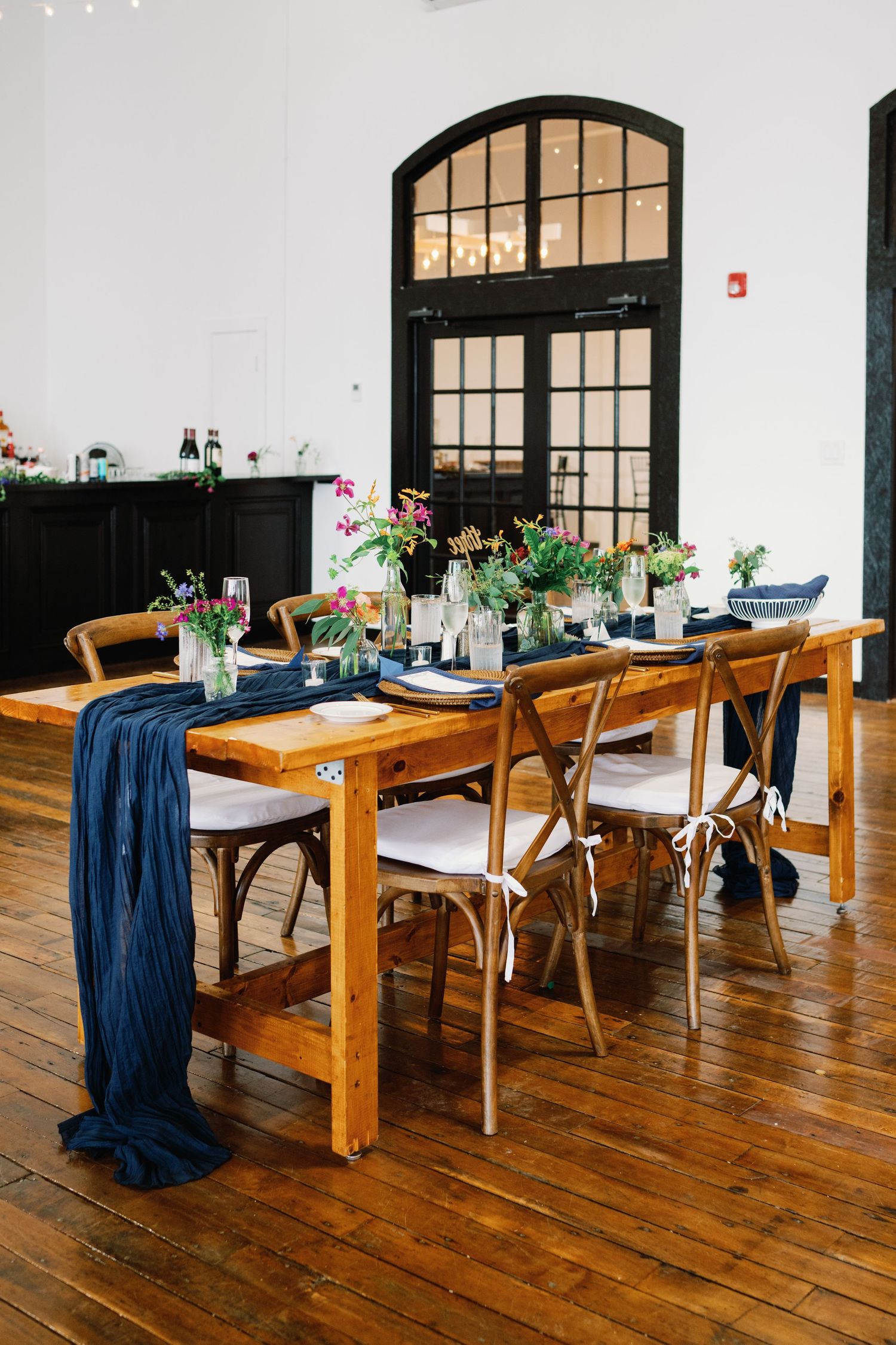 Rustic wooden dining tables with navy blue runners and small floral centerpieces arranged in a row on hardwood flooring.