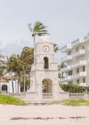 Stone clock tower structure on a beach with palm trees and white buildings in the background.