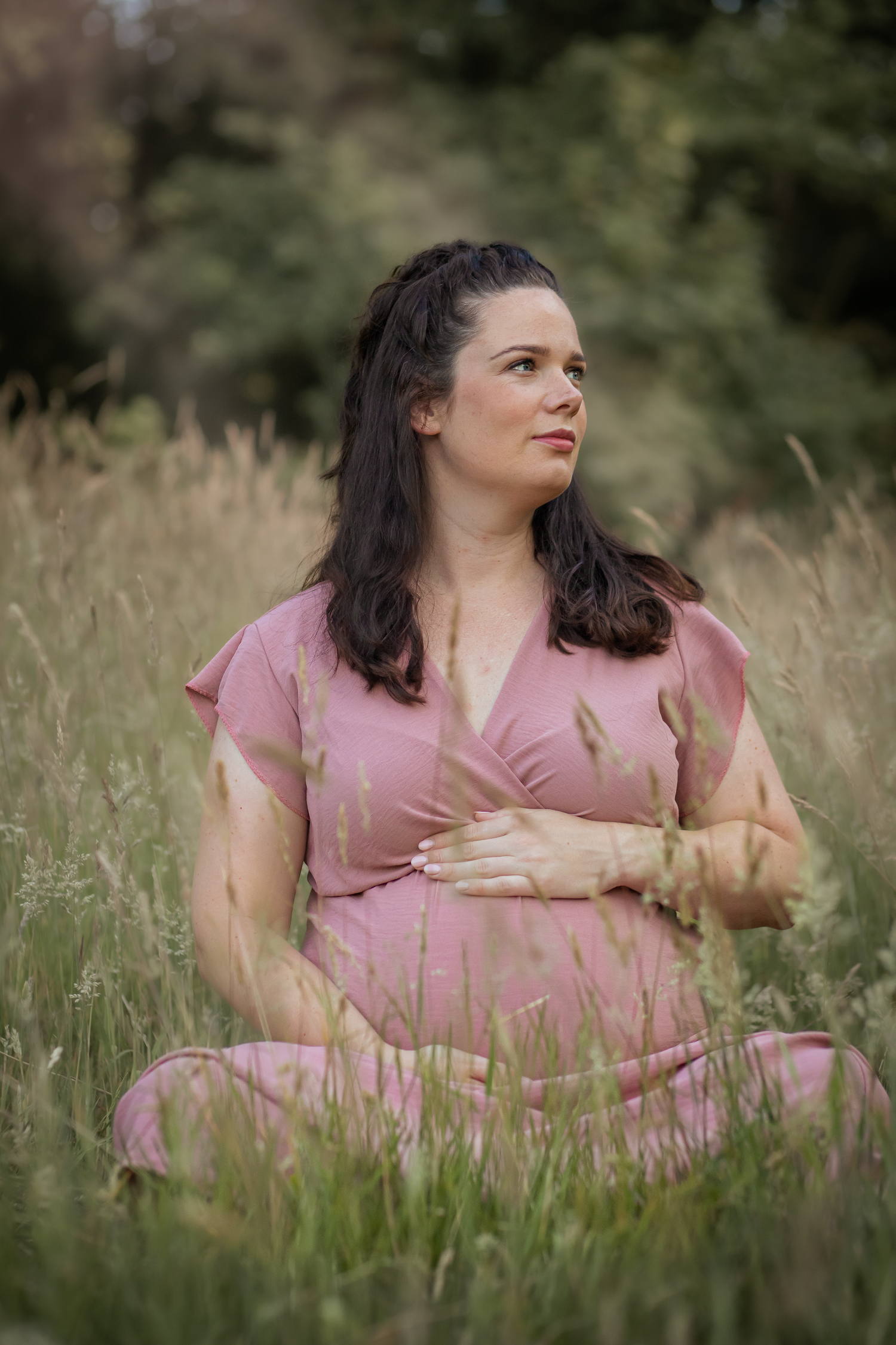Zwangere vrouw in roze jurk zit in een grasveld, dromerige blik opzij.