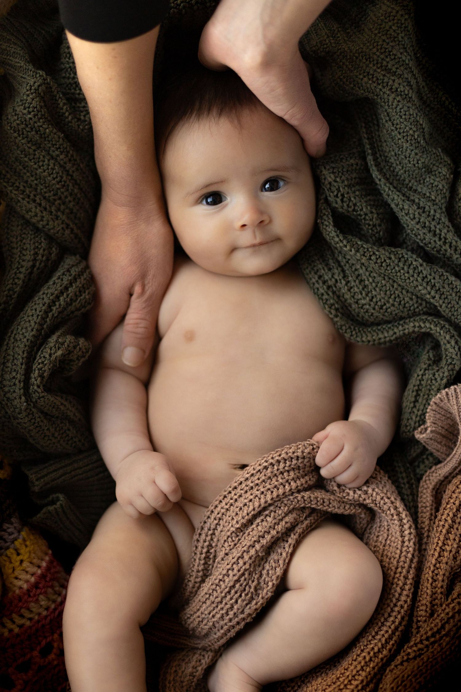 A baby lies on a dark knitted blanket while being gently held by adult hands in a tender moment.