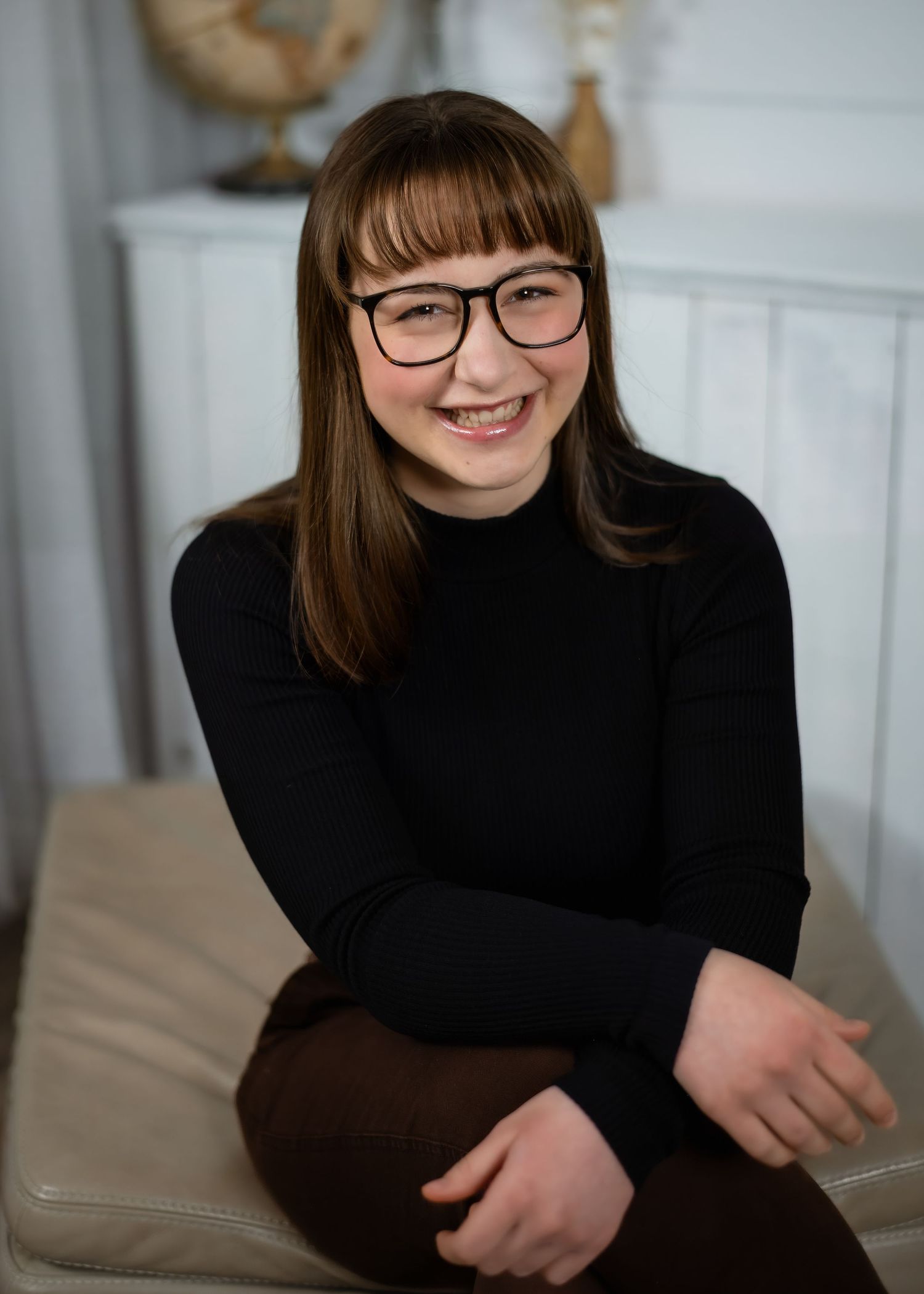 Professional headshot portrait of a person with long brown hair and natural makeup wearing a black off-shoulder top against gray background.
