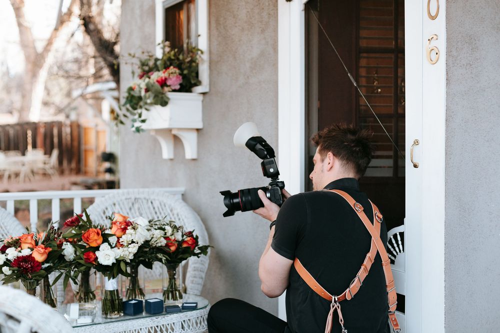 A photographer wearing a black shirt and leather camera strap captures floral arrangements on a white table.