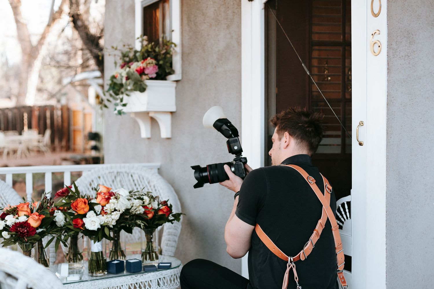 A photographer wearing a black shirt and leather camera strap captures floral arrangements on a white table.