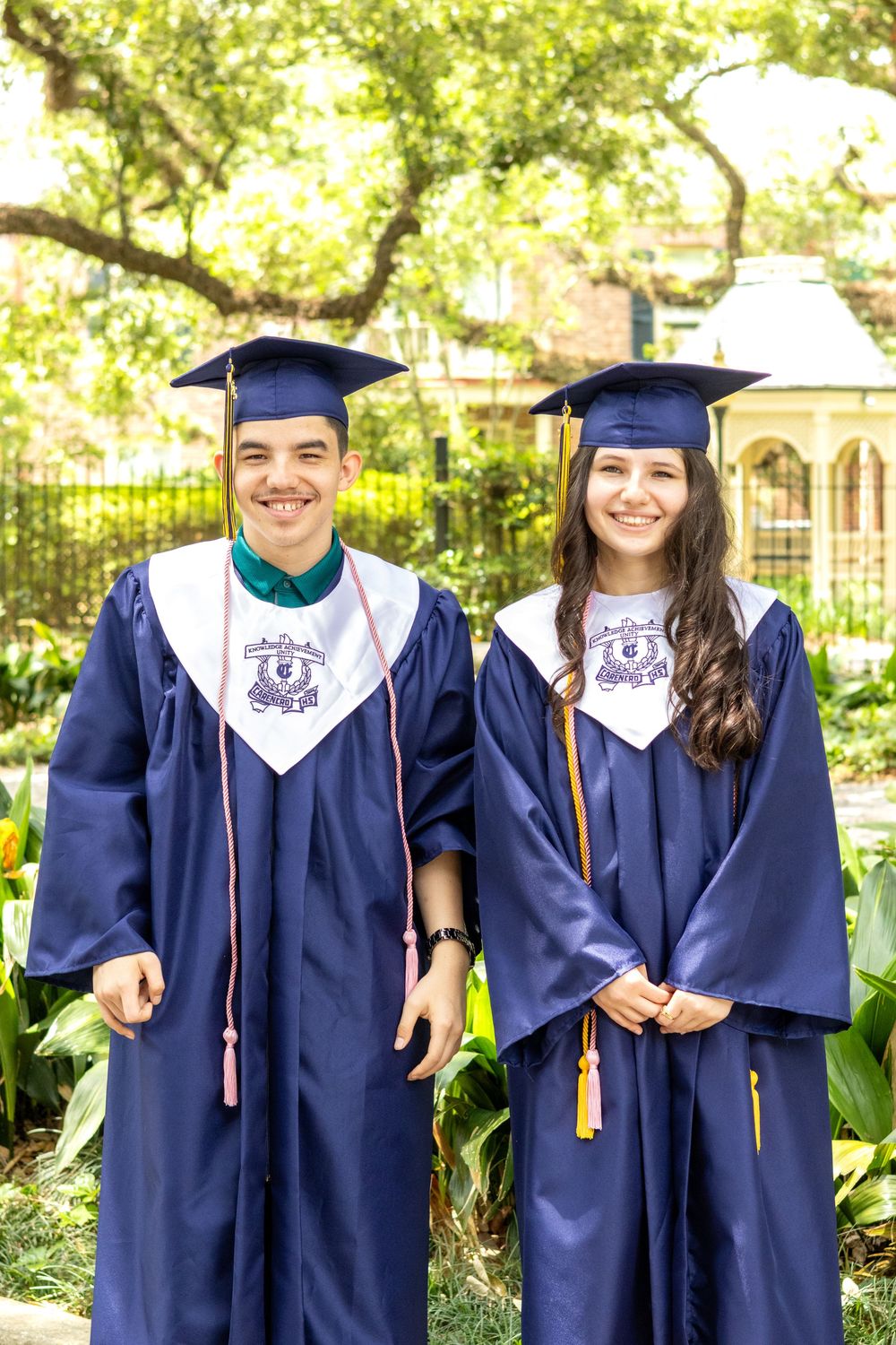 Two graduates in navy blue caps and gowns smile while posing together in a garden setting.