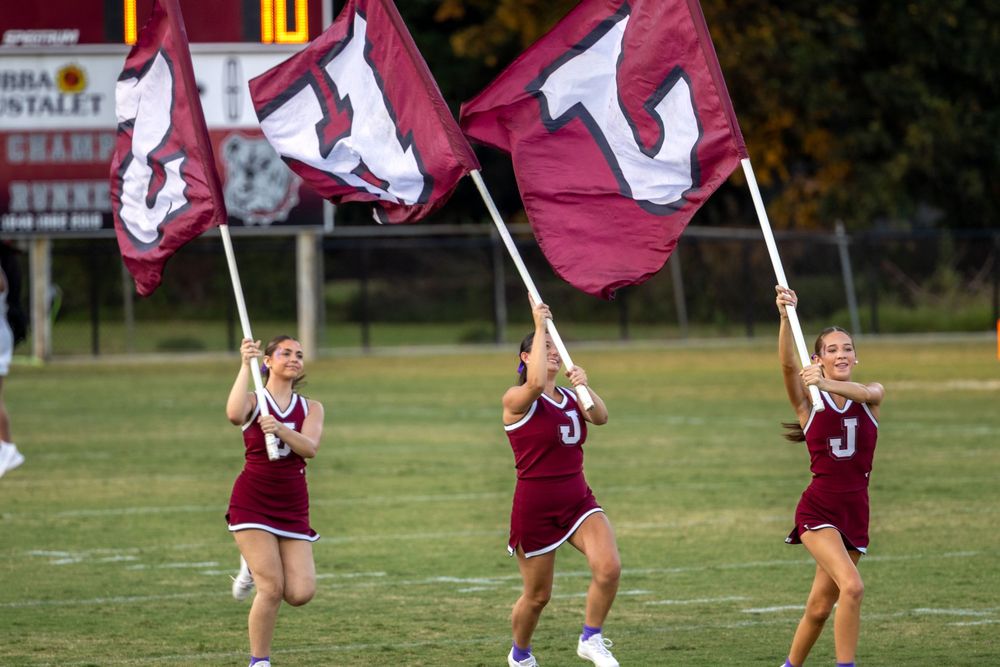 Color guard performers in maroon uniforms practice flag routines on a grassy field during a sunny day.