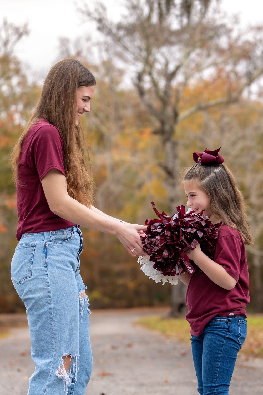 Two cheerleaders in burgundy shirts and jeans practice routines with pom poms in an autumn setting.