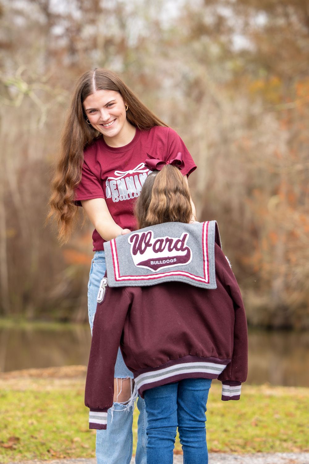 A person in a burgundy sweatshirt and jeans stands outdoors near a lake during autumn.