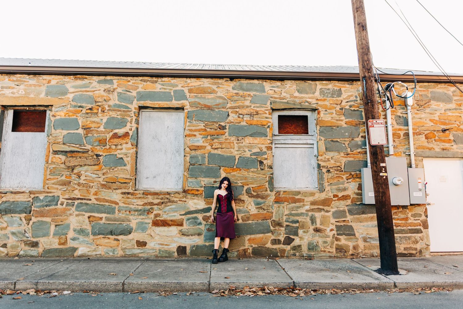 Someone in a purple dress stands against a rustic stone wall of an old building on a city street.