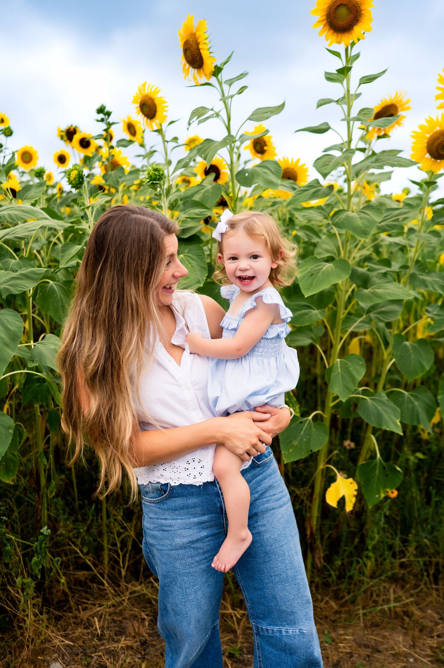 mother smiling looking at toddler daughter in on her hip amidst mammoth sunflowers towering over their heads during mommy and me photoshoot