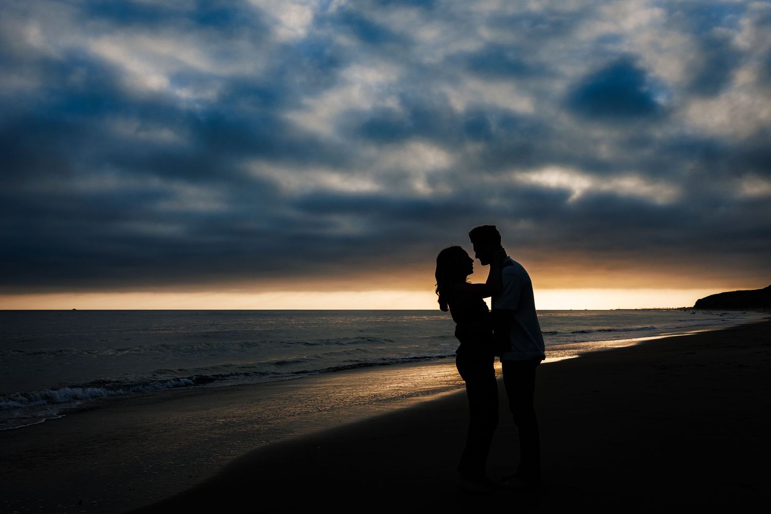 Dark silhouettes of two figures stand together against dramatic sunset sky on beach shoreline.