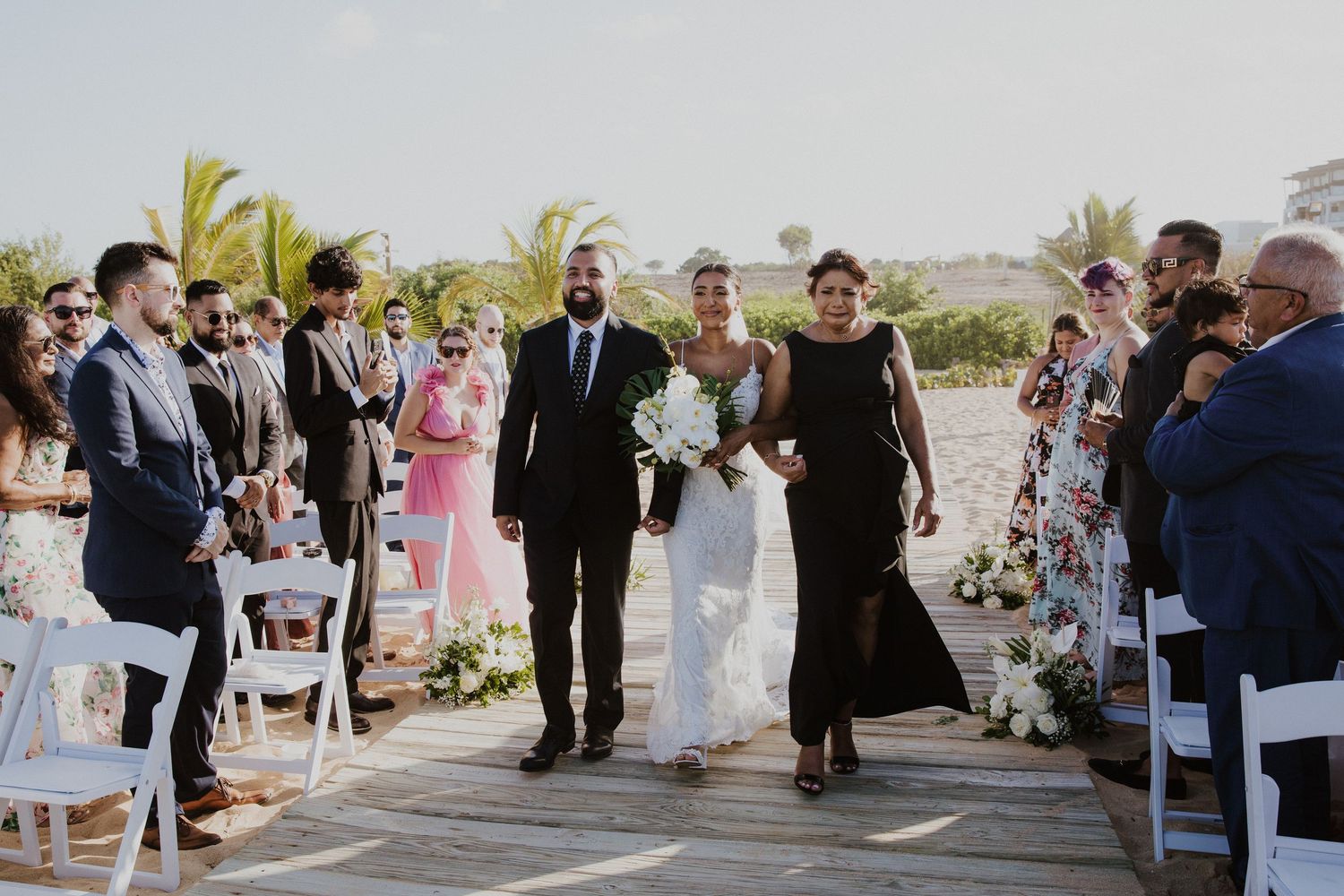 A beach wedding ceremony procession on a wooden boardwalk lined with white chairs and tropical palm trees.