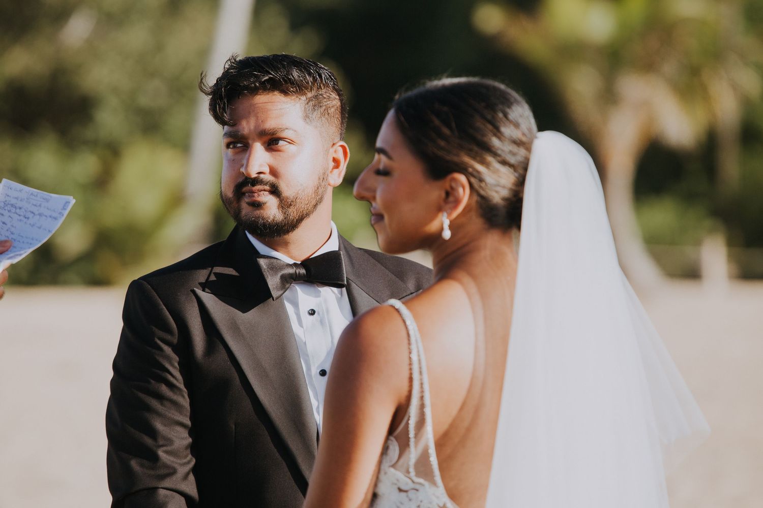 A close-up moment during an outdoor wedding ceremony with a white floral arch in the background.