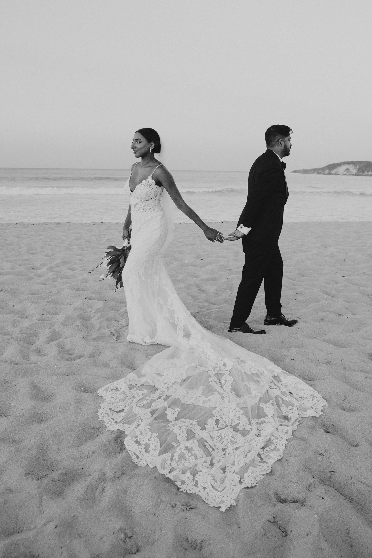 Dramatic beach wedding photo of couple walking with bride's lace train flowing behind them.