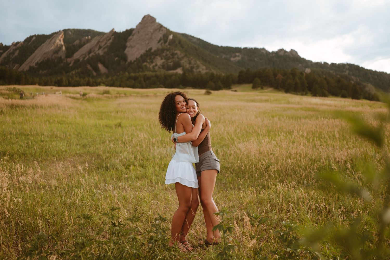 Sisters embrace in a grassy meadow with the Flatirons mountain formation rising dramatically in the background near Boulder, Colorado.