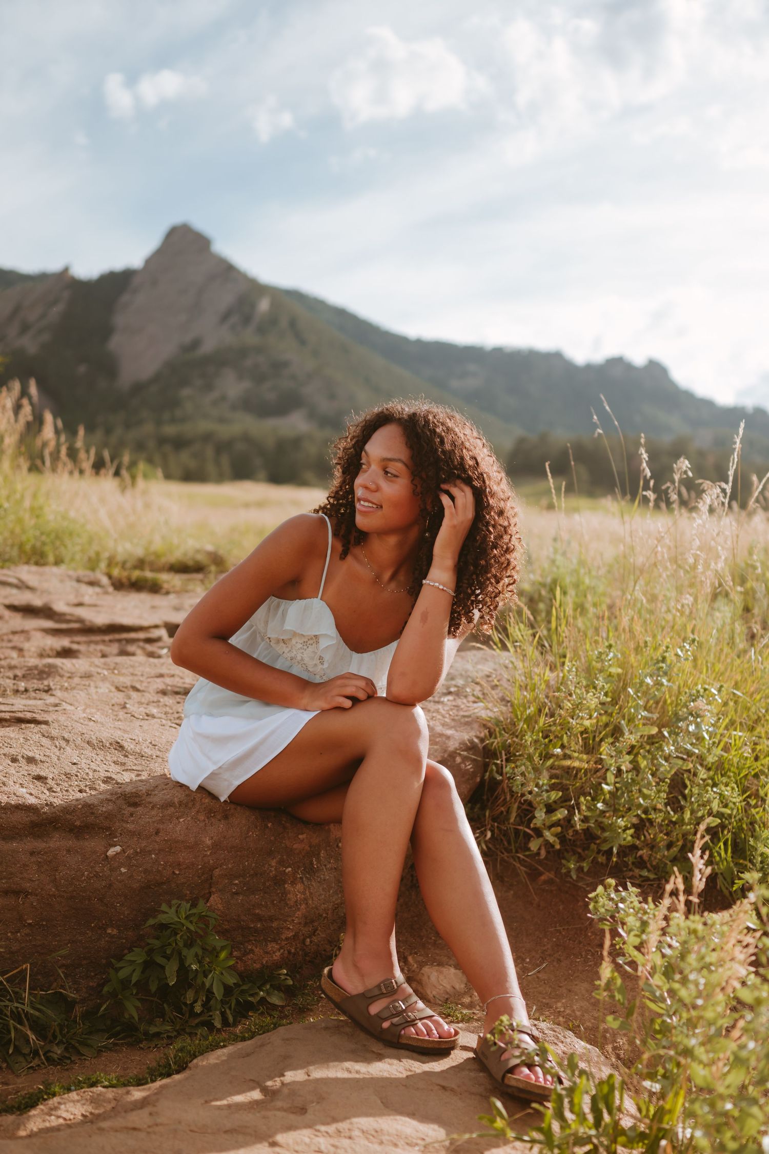 Woman in white dress sits on mountain trail during golden hour with scenic peaks in background.