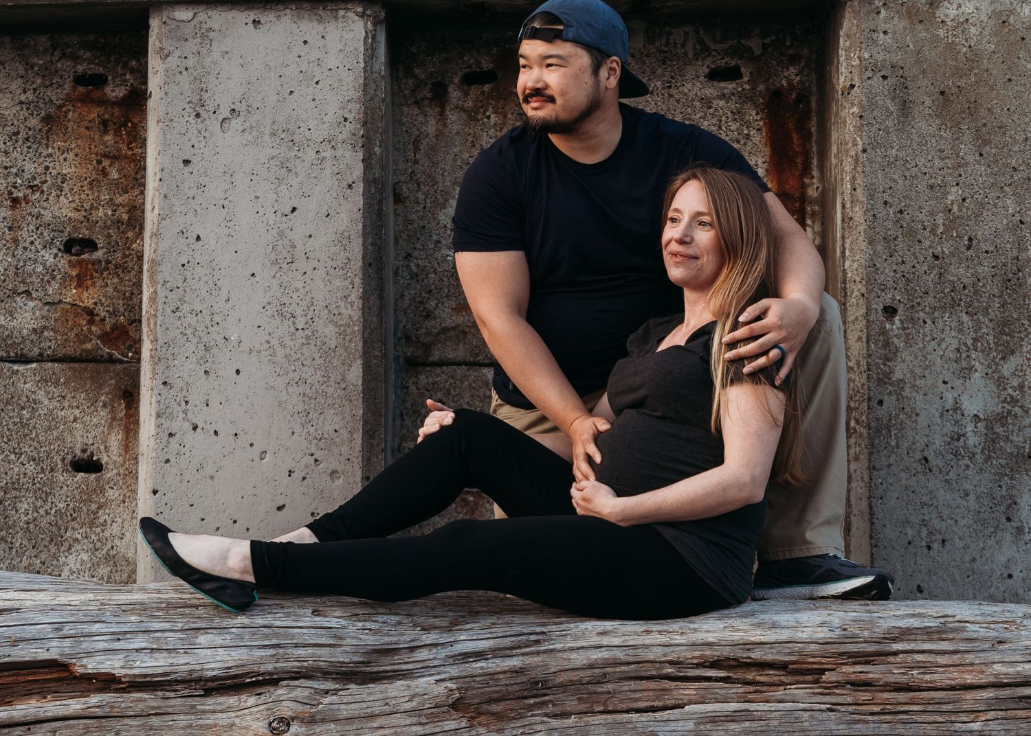 A couple dressed in black sits together and shares a laugh while resting against a concrete column outdoors.