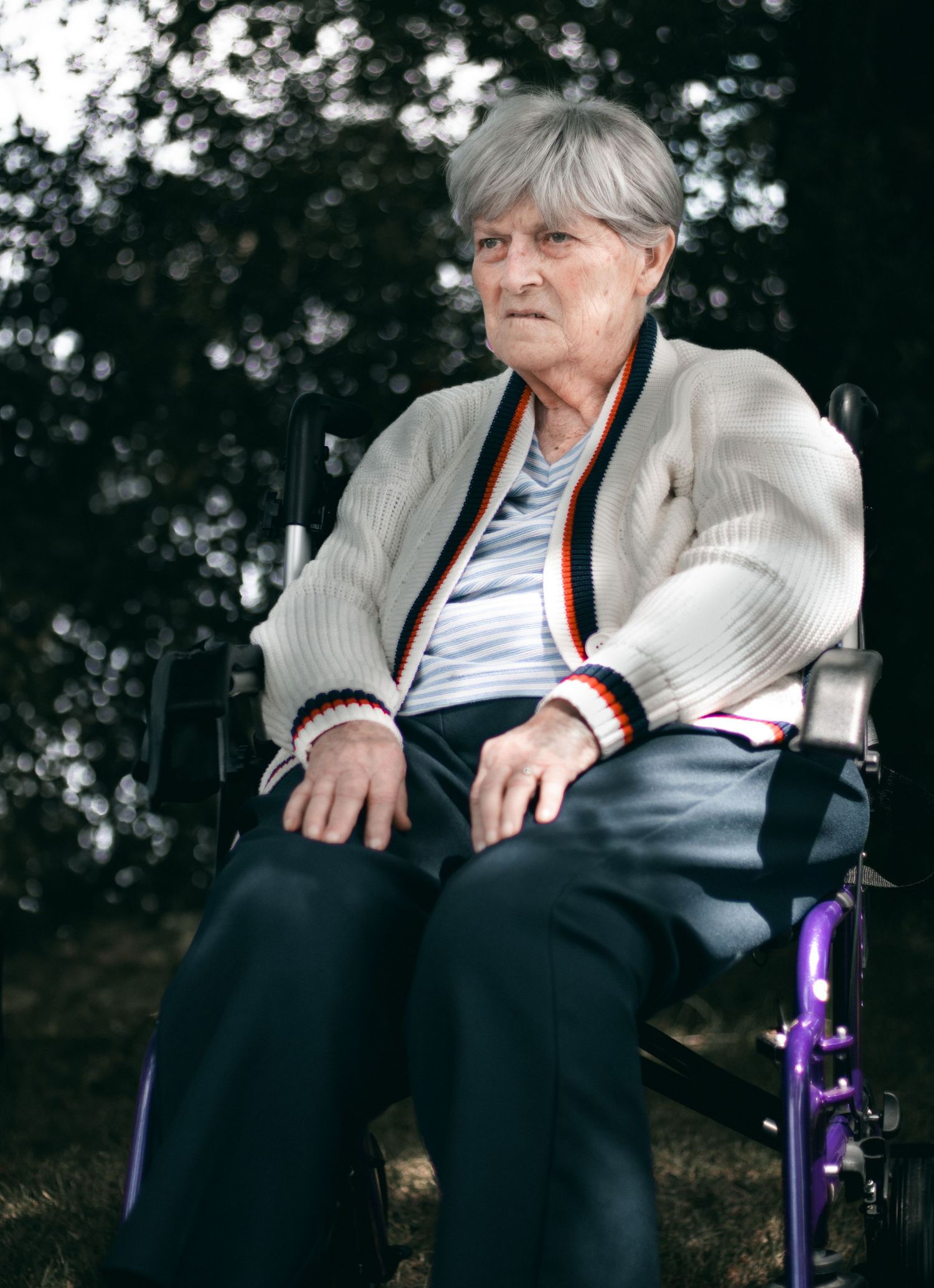 Person seated in a purple wheelchair wearing a white cardigan and striped shirt outdoors.