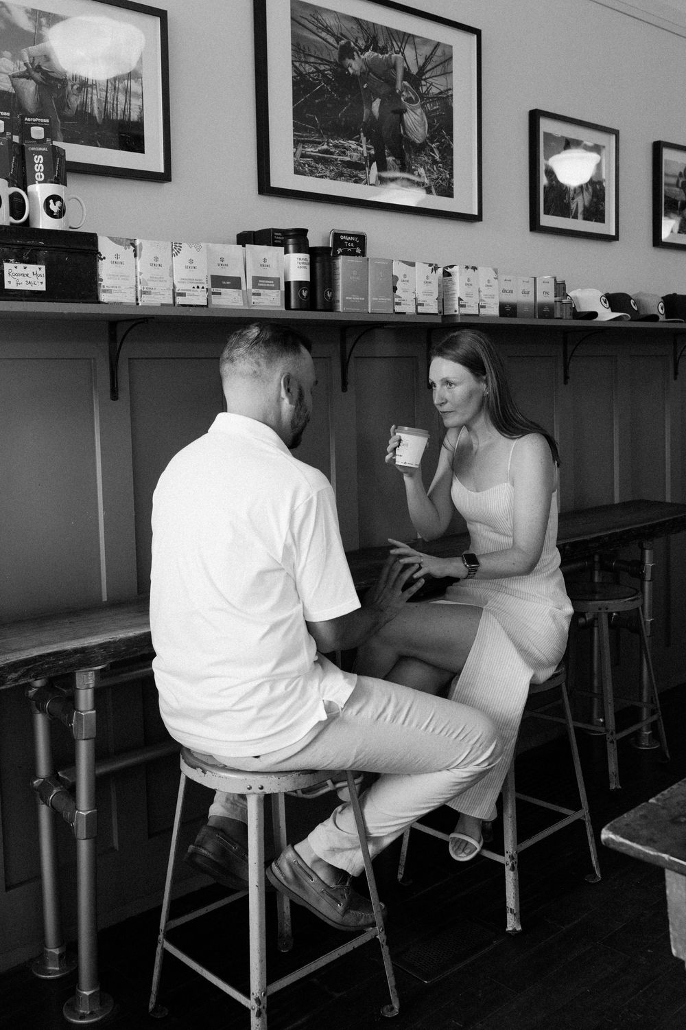 Two people enjoy coffee while sitting on bar stools at a cafe with black and white photos on the wall.