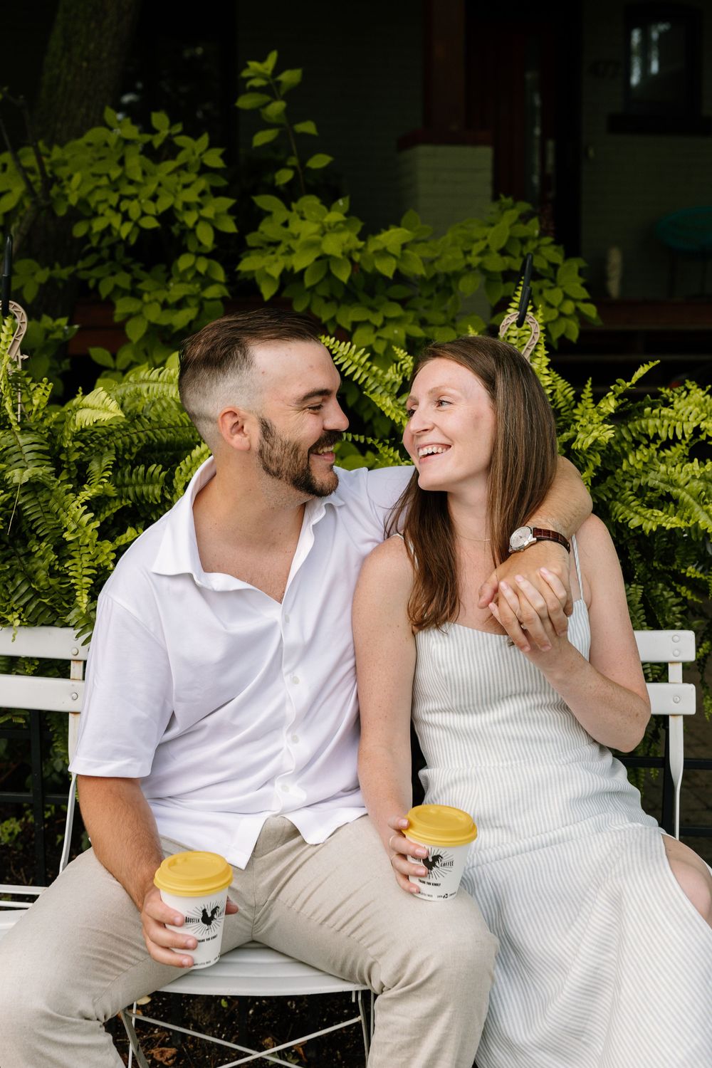 A couple sharing a joyful moment while sitting on a white bench surrounded by lush ferns.