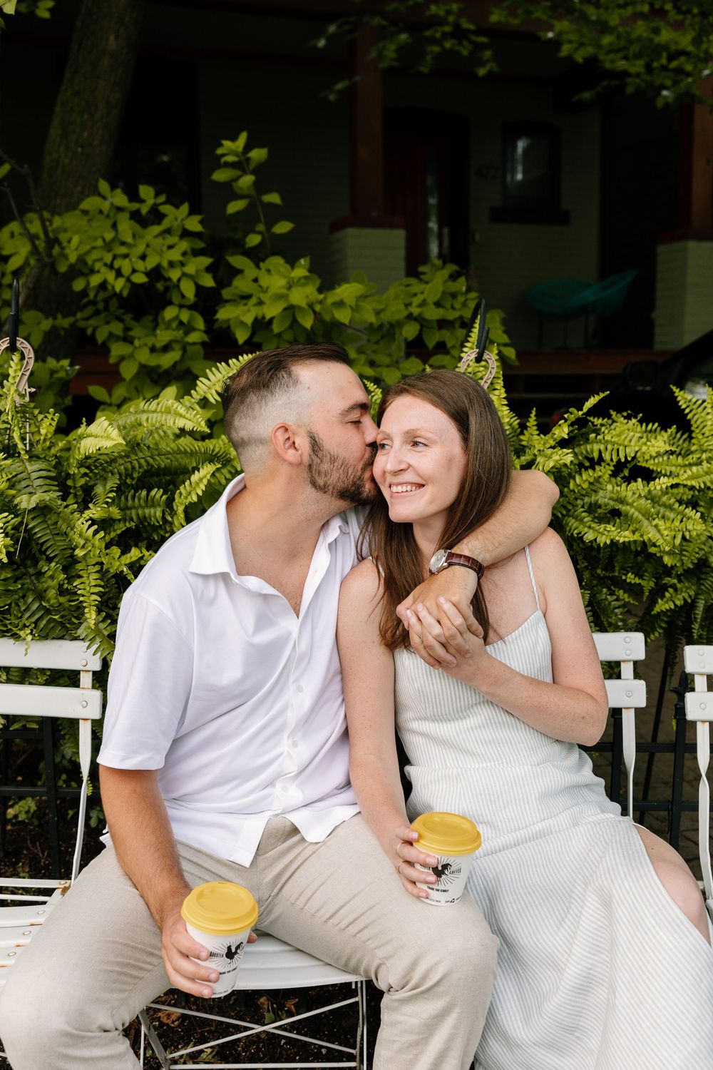 A couple shares a sweet romantic moment while sitting on white chairs in a lush garden setting.