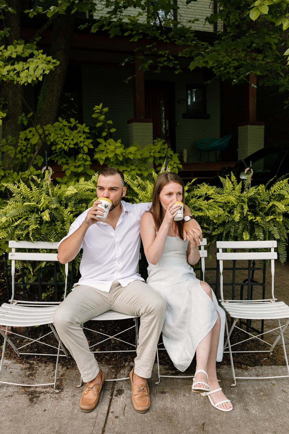 A couple sits on white folding chairs outdoors while enjoying beverages in a garden setting.