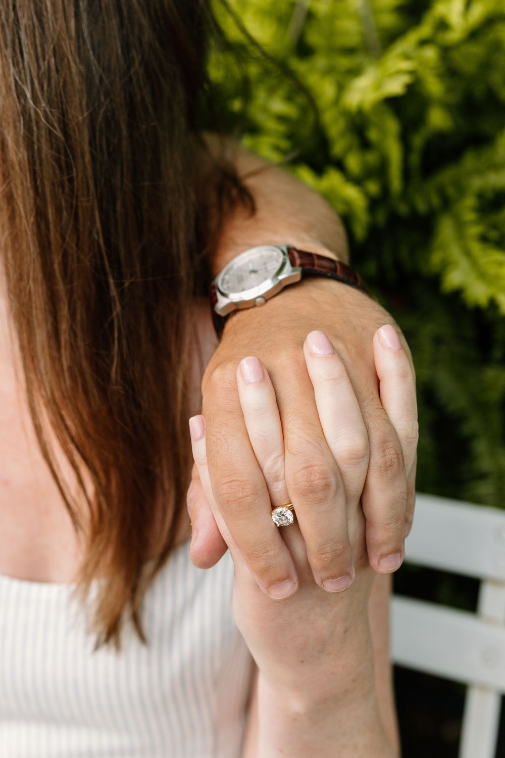 A delicate engagement ring sparkles on a hand, with a wristwatch and green foliage in the background.