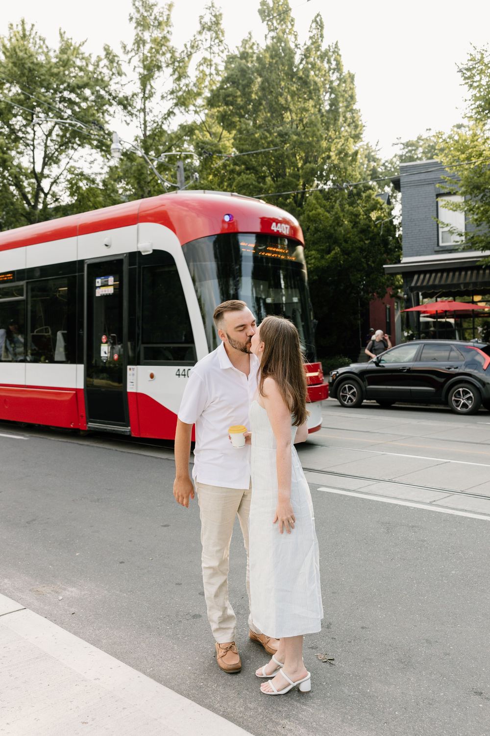 A couple in light summer outfits share a romantic moment on a city street with a red streetcar in the background.