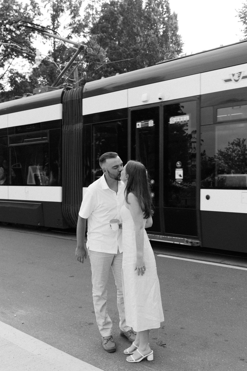 A couple in white outfits sharing an intimate moment in front of a modern tram on a sunny day.