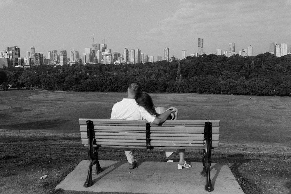 Person sitting alone on park bench overlooking Toronto city skyline through trees.