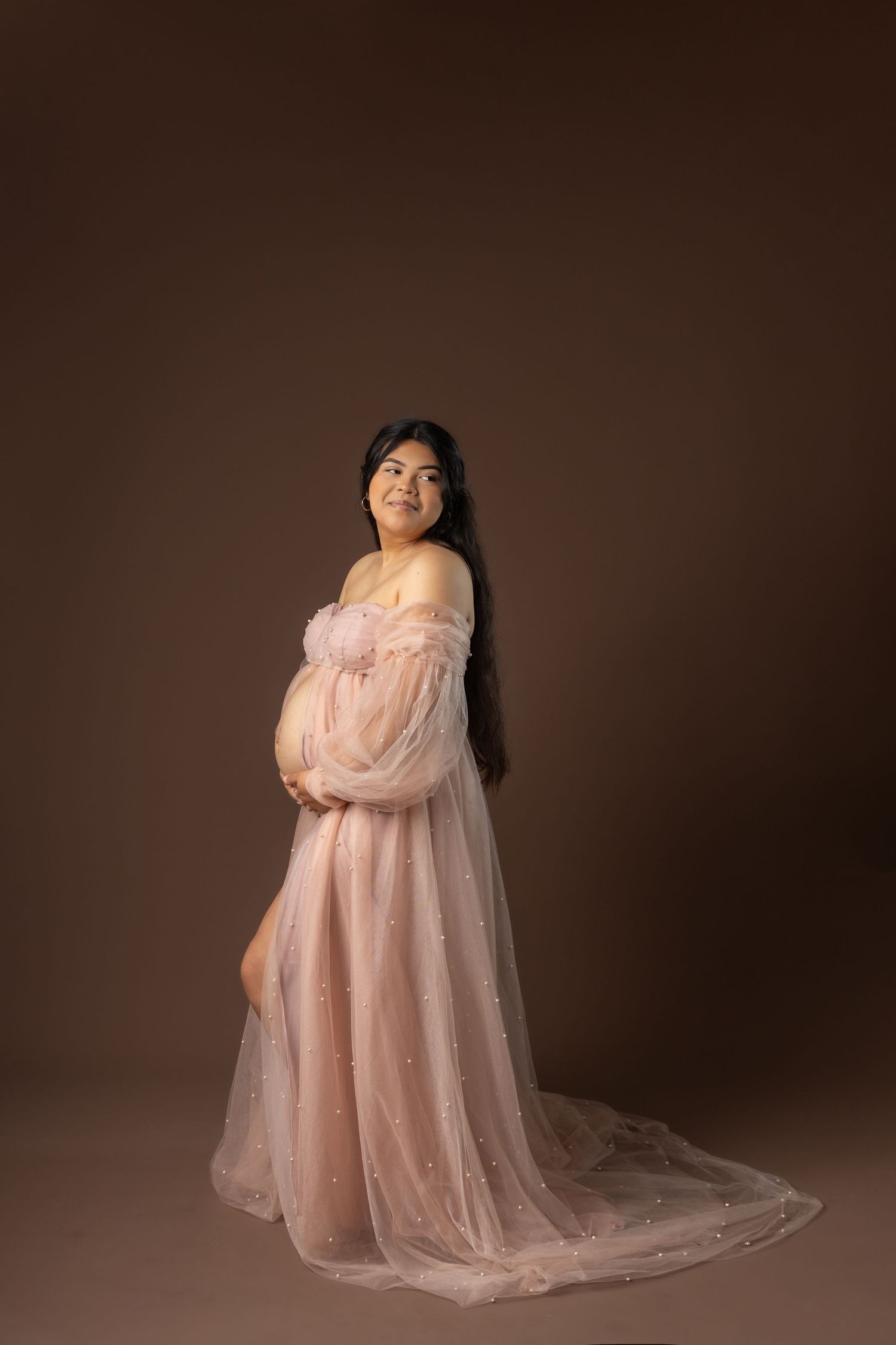 An elegant maternity portrait in a flowing blush pink tulle gown with sheer sleeves against a dark gray backdrop.