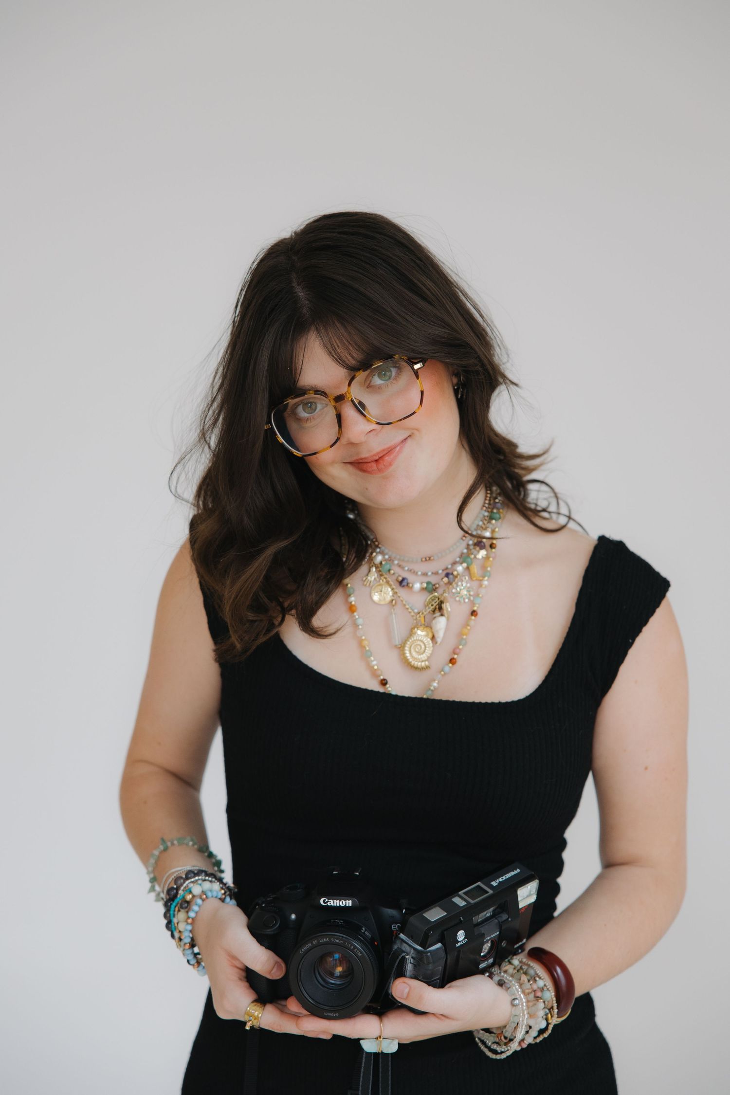 Photographer in a black top and denim holds professional camera while wearing stylish glasses and jewelry against white wall.